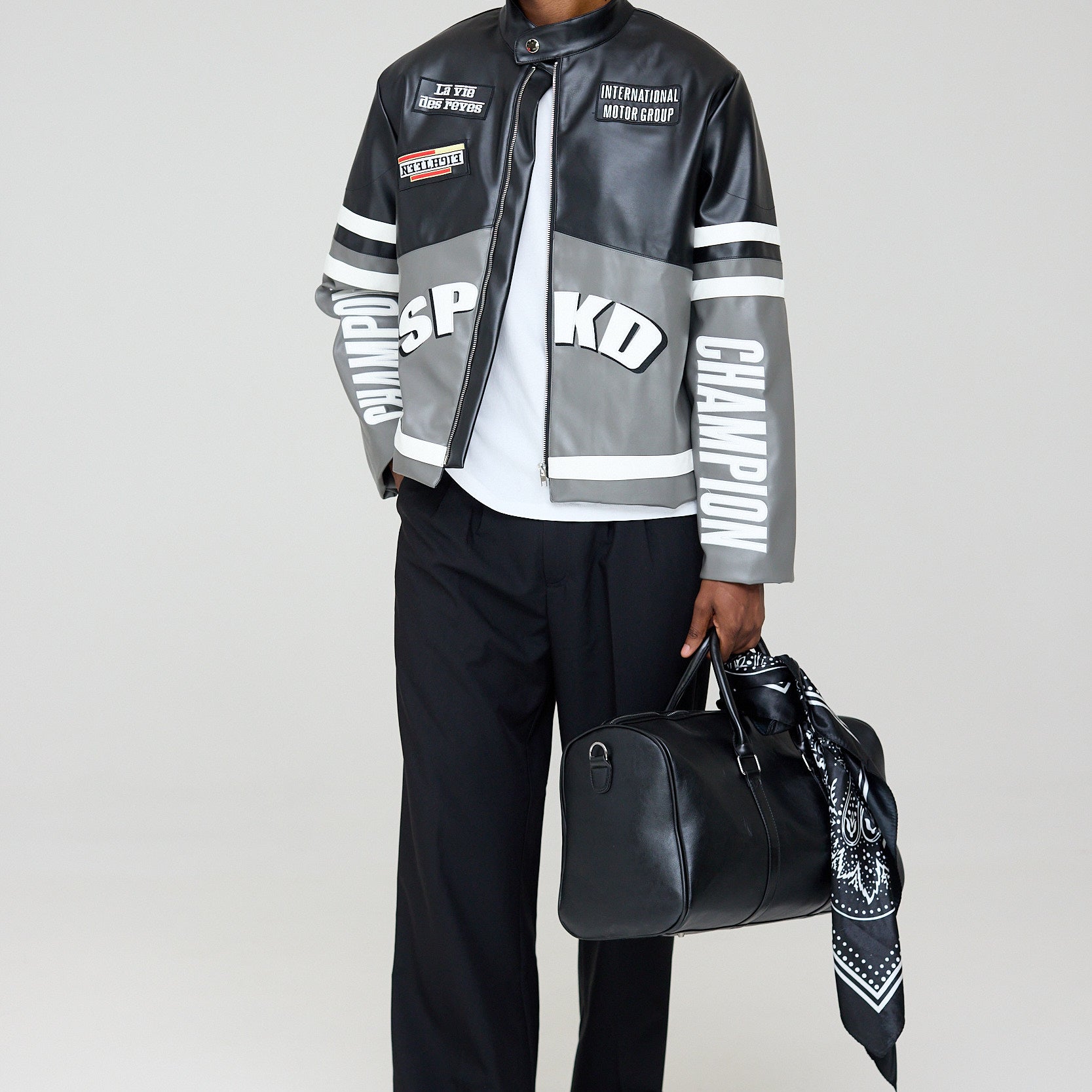 Man wearing a stylish jacket with brand logos, holding a black bag on a white background