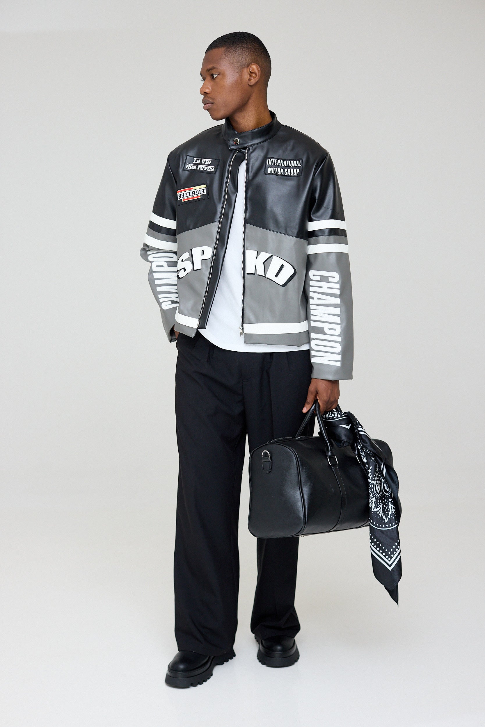 Man wearing a stylish jacket with brand logos, holding a black bag on a white background