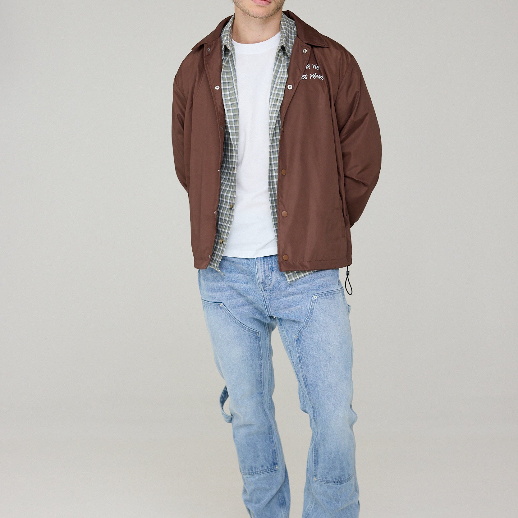 Man wearing a brown jacket, white shirt, plaid shirt, and blue jeans on a plain background