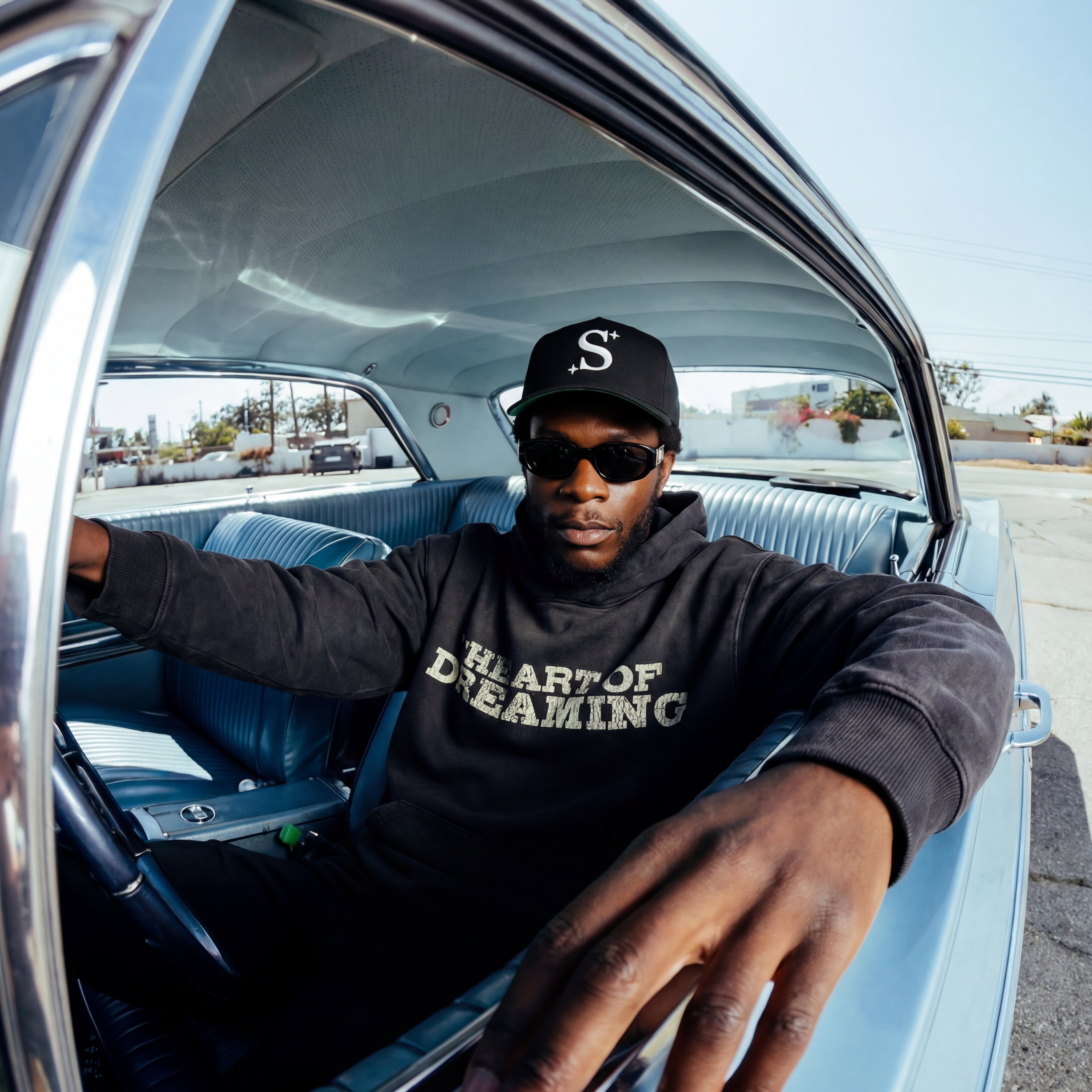 Person sitting inside a classic car wearing a black hoodie with 'The art of dreaming' text along with a S trucker cap.