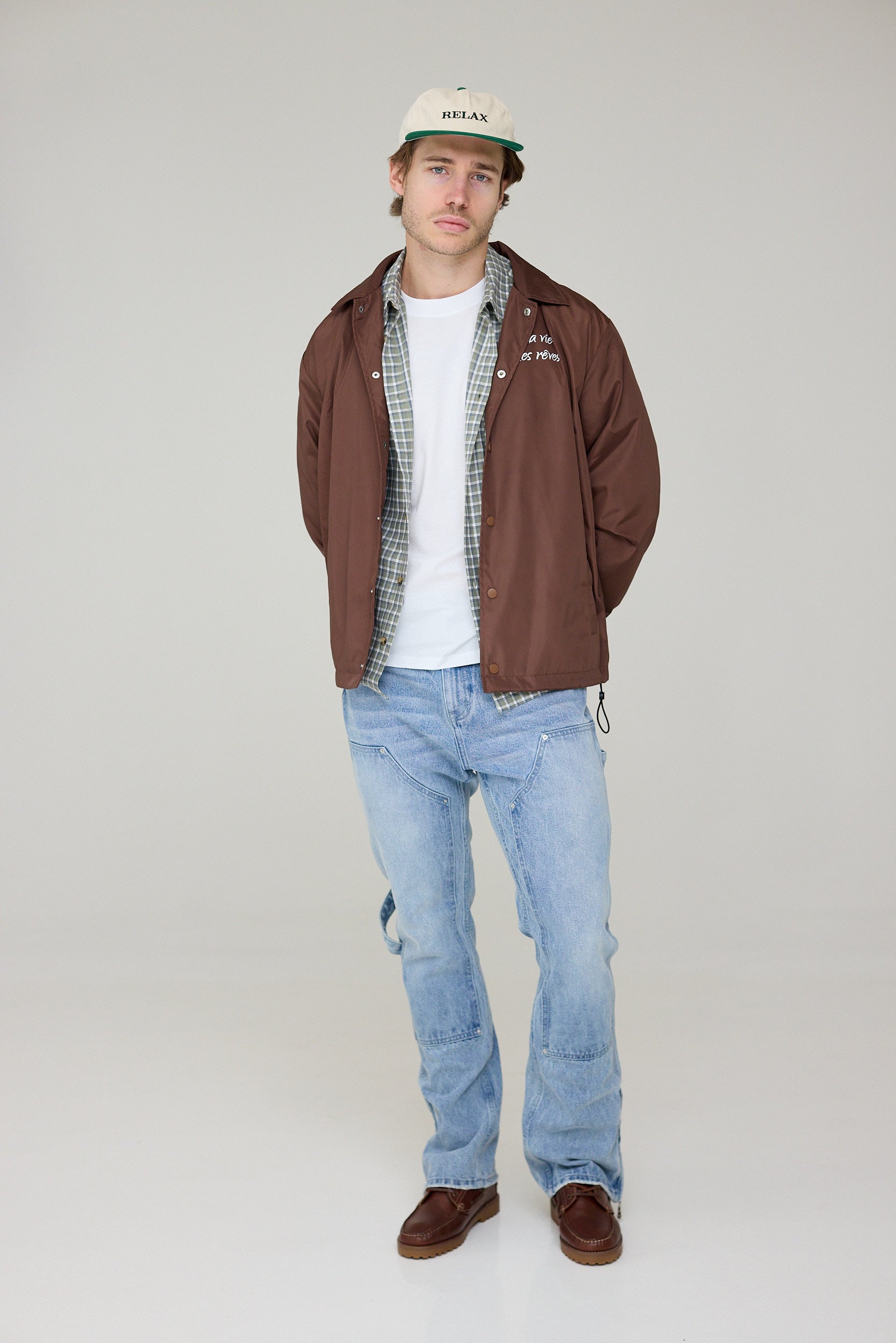 Man wearing a brown jacket, white shirt, plaid shirt, and blue jeans on a plain background